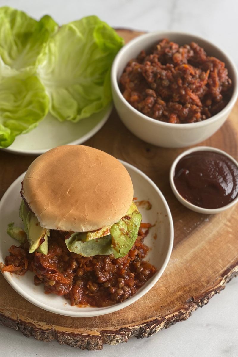 vegan sloppy joe on a plate on a wood cutting board next to a plate of lettuce leaves and bowls of more filling and BBQ sauce