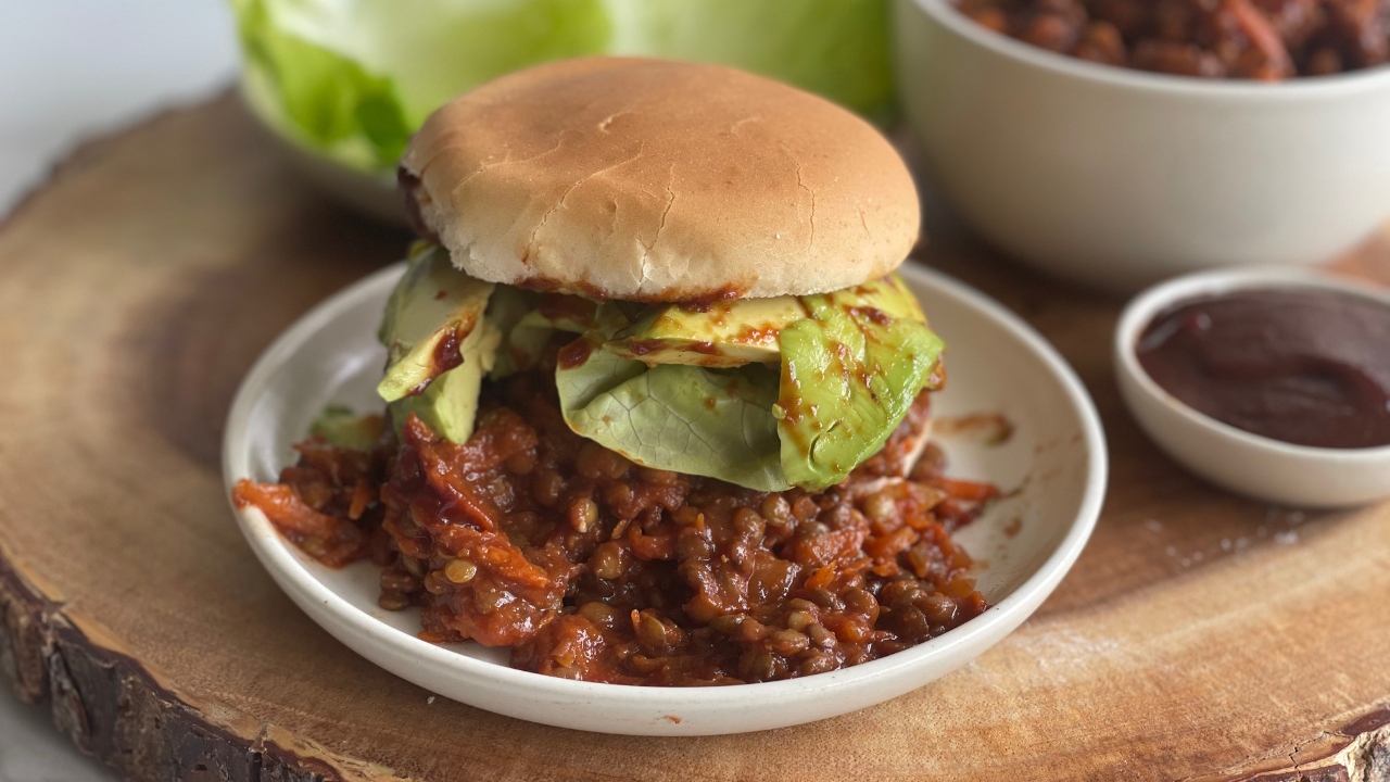 sloppy joe on a plate on a wood cutting board next to bowls of barbecue sauce and more sloppy joe filling