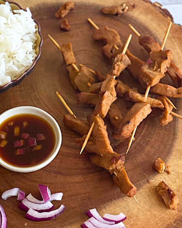 3 seitan skewers on a wood cutting board next to a bowl of rice and a bowl of teriyaki sauce