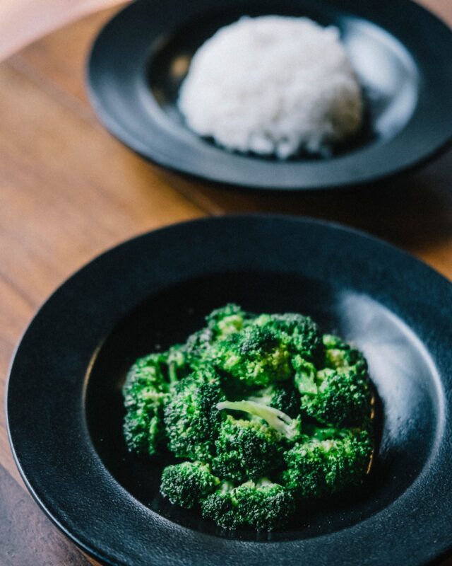 A bowl of steamed broccoli on a table in front of a bowl of white rice