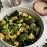 Bowl of broccoli and tempeh on a counter next to a wooden bowl of dressing with a spoon in it