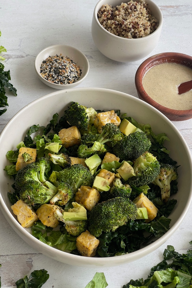 bowl of tempeh and broccoli on a counter next to a bowl of Everything seasoning and a wooden bowl of dressing with a spoon in it