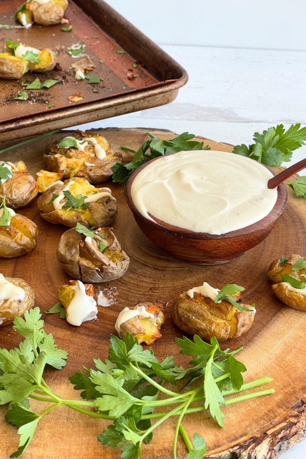 cutting board with smashed potatoes on them next to a wooden bowl filled with truffle cream. A sheet pan with more potatoes sits behind and above it.