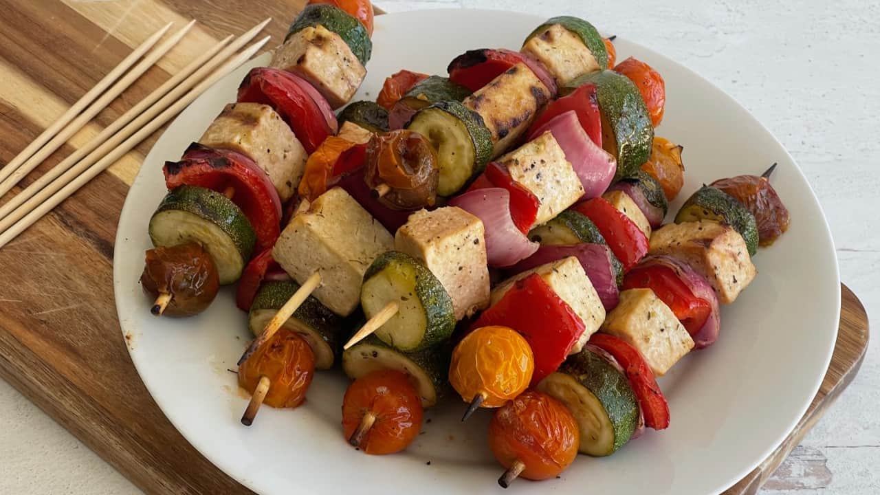 6 skewers of tofu and vegetables on a white plate next to several wood skewers on the left
