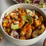 bowl of tofu and cauliflower drizzled with Buffalo sauce surrounded by bowls of Buffalo sauce, avocado and coleslaw