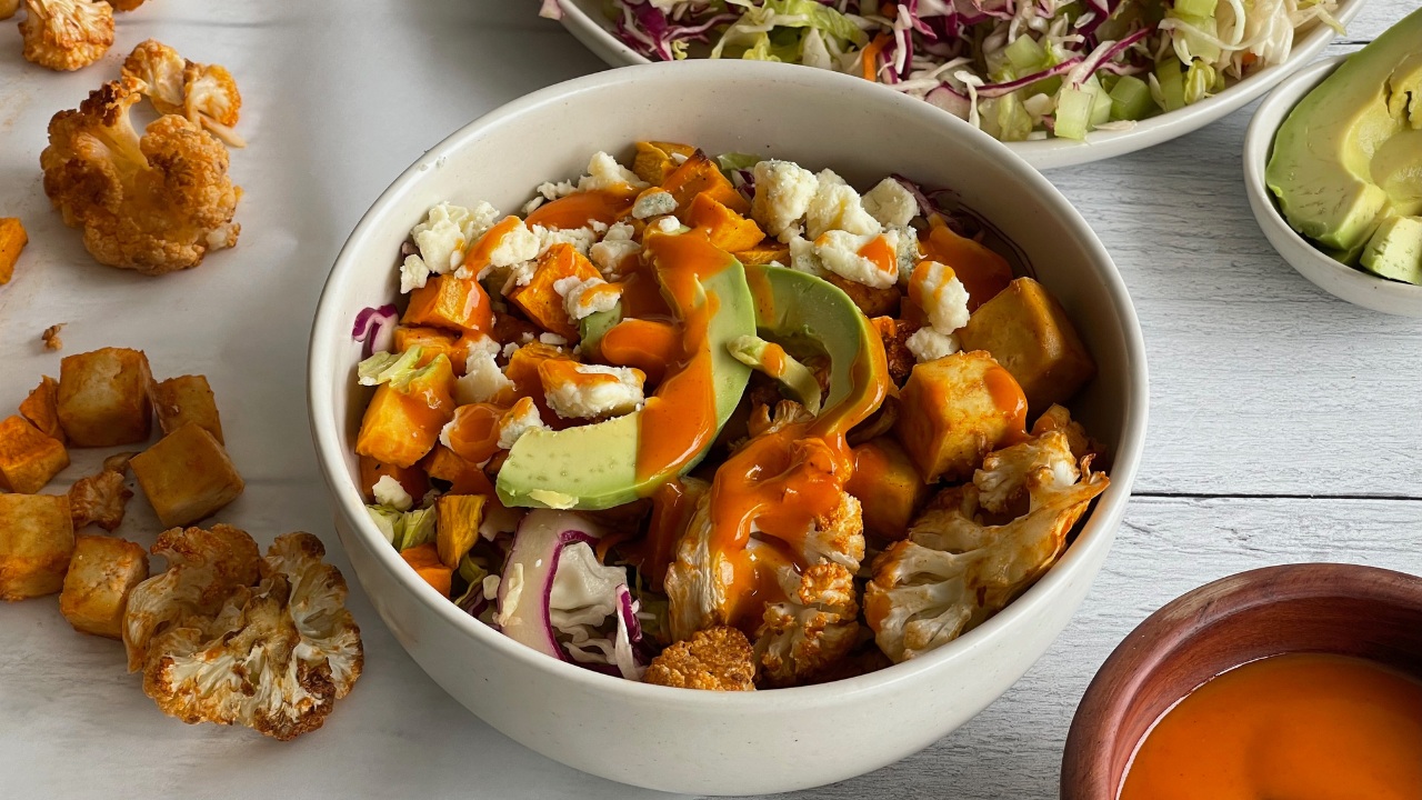 bowl of cubed tofu and sweet potatoes and cauliflower florets topped with Buffalo sauce, avocado and bleu cheese
