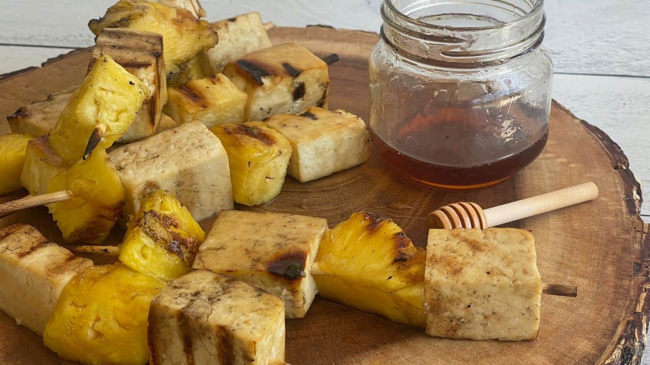 Skewers of grilled pineapple and tofu on a cutting board alongside a jar of honey with a honey stick lying next to it