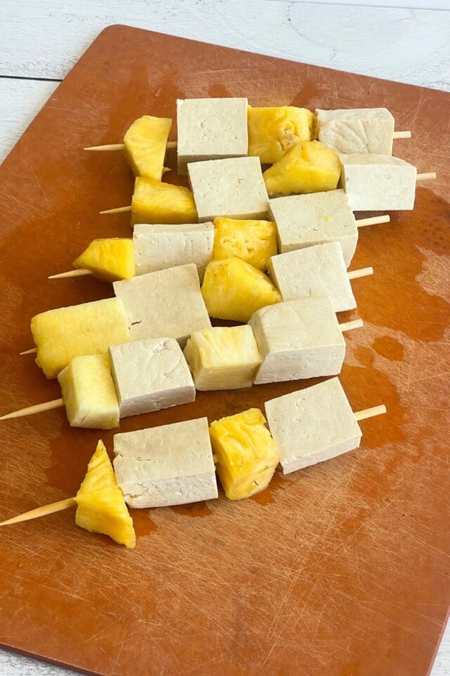 Wood cutting board with five wood skewers threaded with chunks of tofu and pineapple