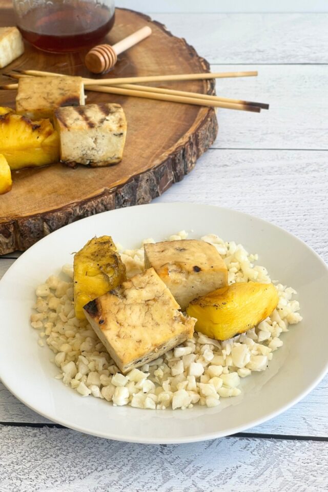 plate of cauliflower rice with chunks of grilled tofu and pineapple on top of it, with a wood cutting board behind the plate with kebabs on top