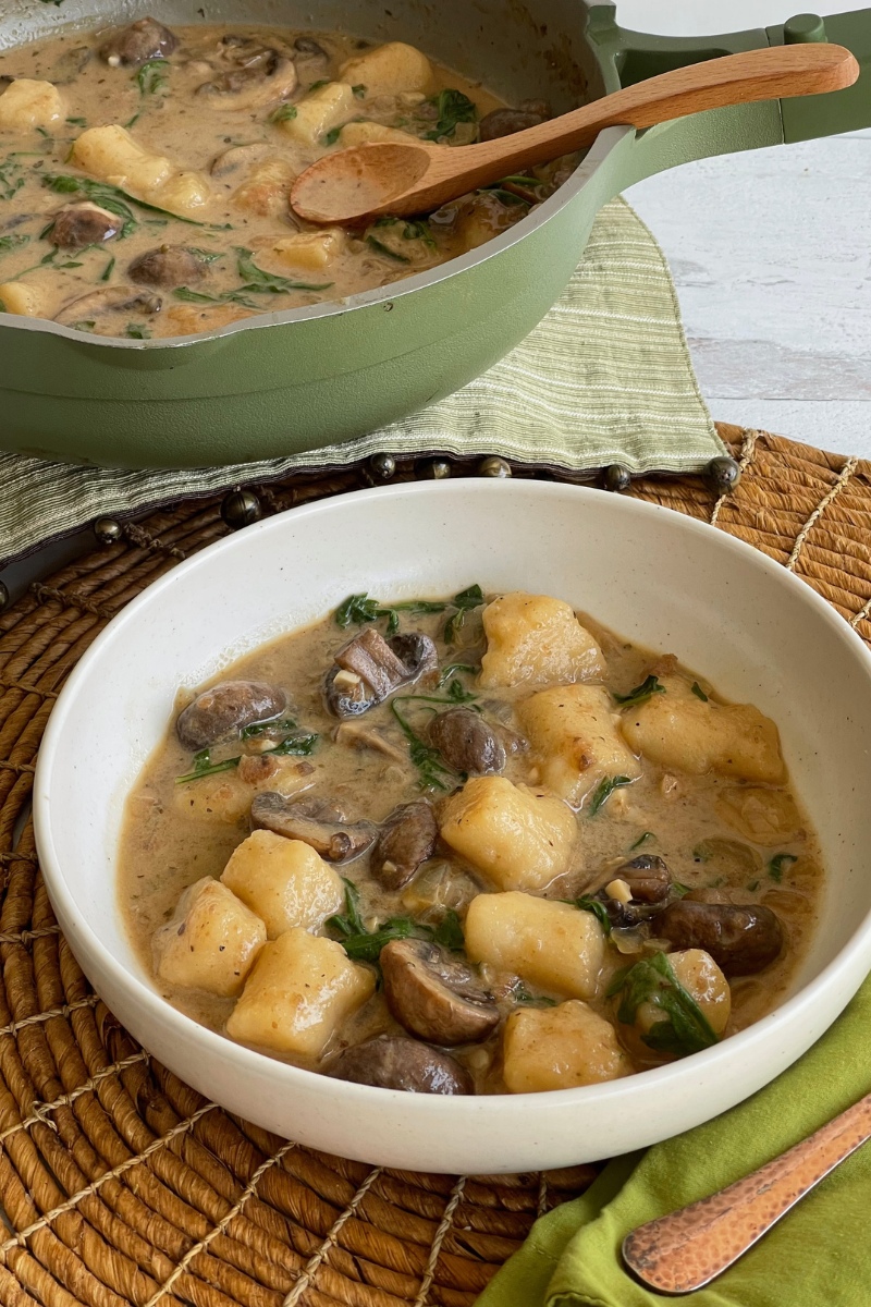bowl of gnocci and mushrooms in sauce in front of a pan with more of the dish with a wooden spoon in it