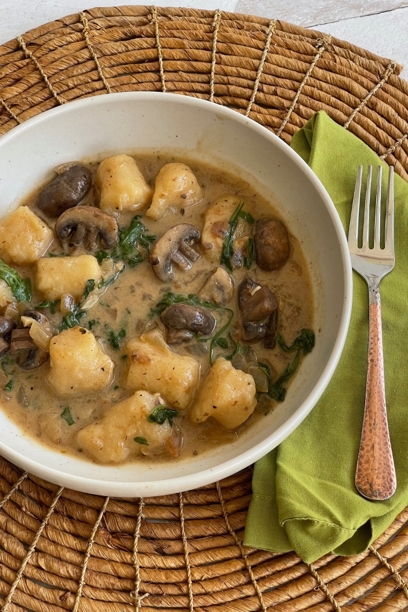 bowl of gnocchi and mushrooms in a sauce on a wicker placemat next to a green napkin with a fork on it