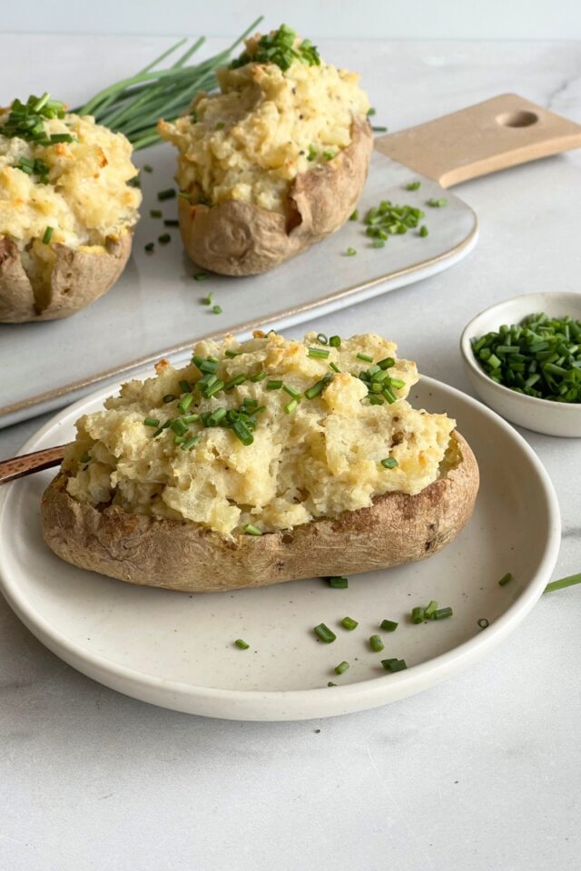Stuffed baked potatoe on a plate in front of a white serving platter with 2 more potatoes
