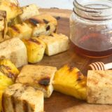 four pineapple and tofu skewers on a wood cutting board next to a jar of hot honey