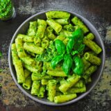 bowl of rigatoni with pesto sauce on a countertop next to a fork and basil leaves