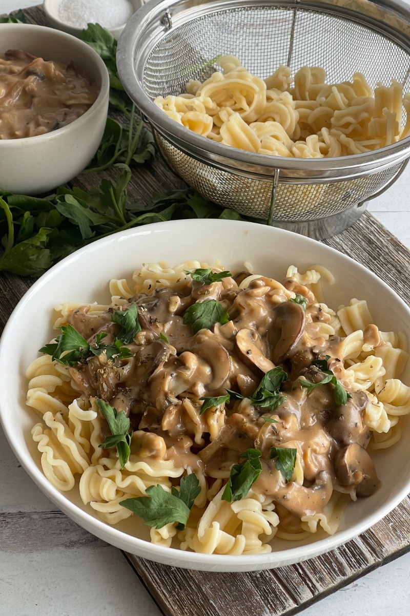 bowl of mushroom stroganoff on a wood board in front of a colander of noodles