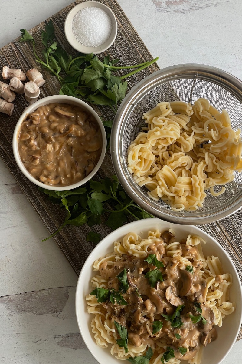 bowl of mushroom stroganoff on a wood bowl in front of a colander of noodles, a bowl of sauce, mushroom stems and a bowl of salt