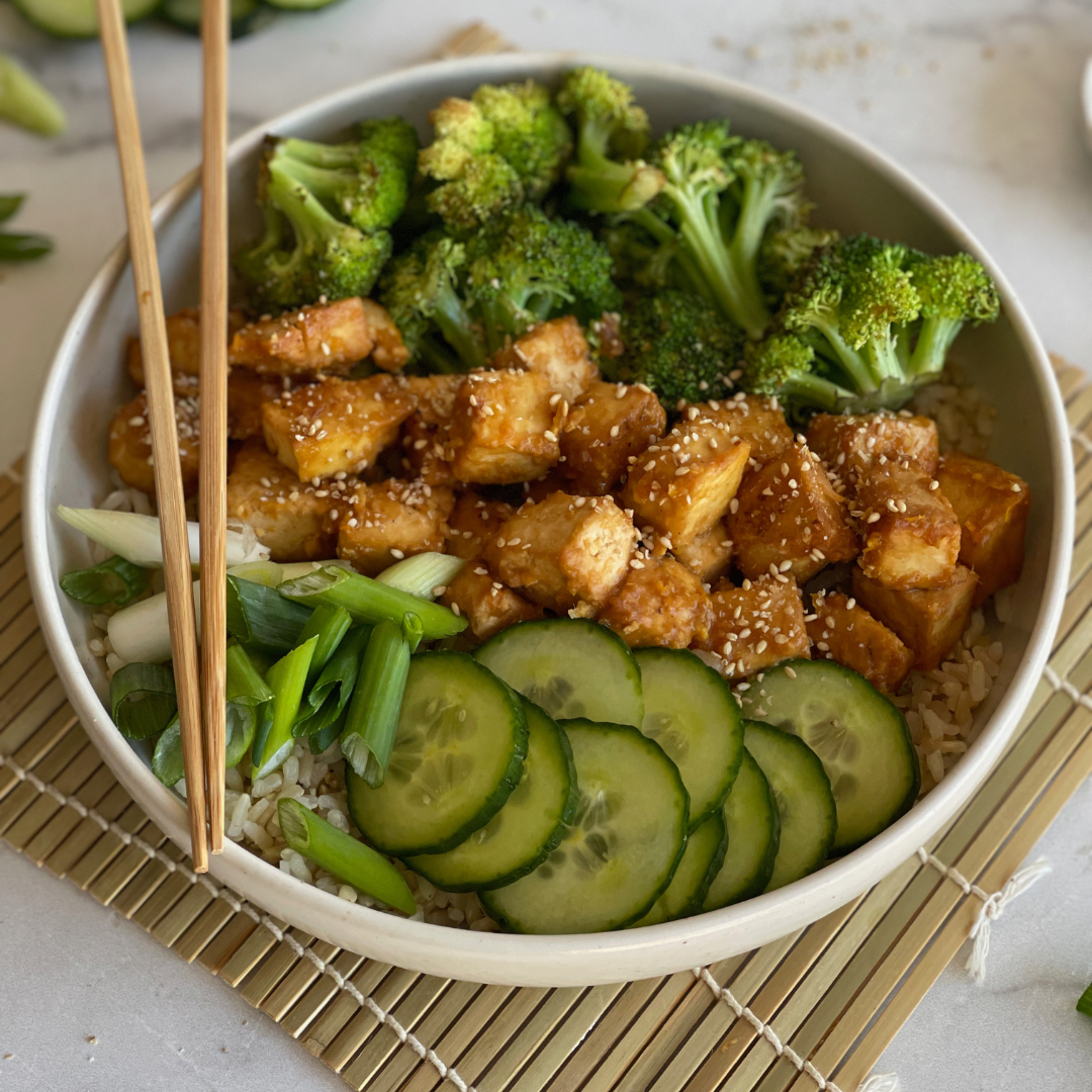 bowl of broccoli, tofu and cucumbers on top of a grass mat