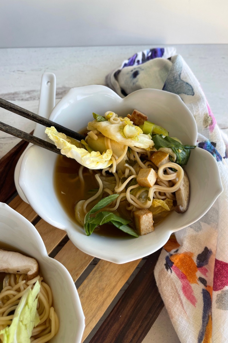 bowl of ramen with chopsticks in it on top of a white floral napkin and next to a white ceramic soup spoon on a wood cutting board