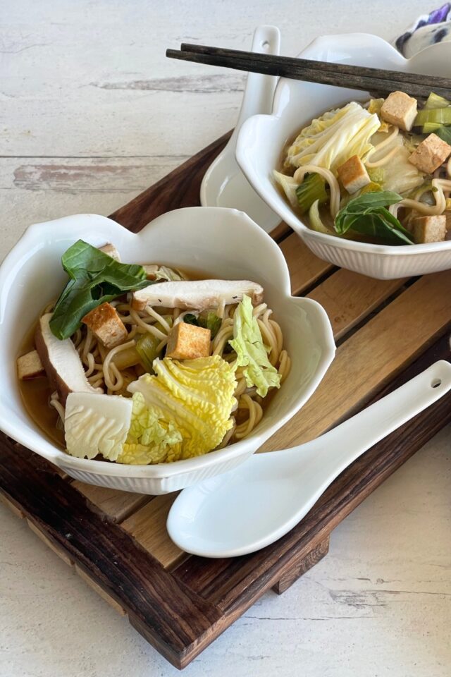 Two bowls of ramen on a wood cutting board with a ceramic white spoon next to one bowl and a set of chopsticks resting on the other bowl