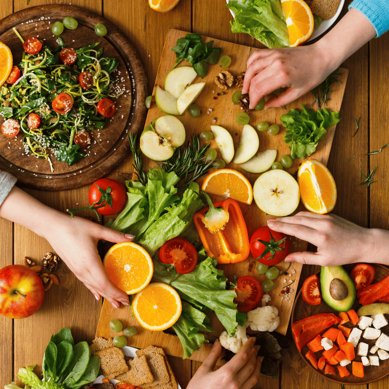 table with a cutting board full of fruits and vegetables next to a salad bowl with 3 hands grabbing for the food