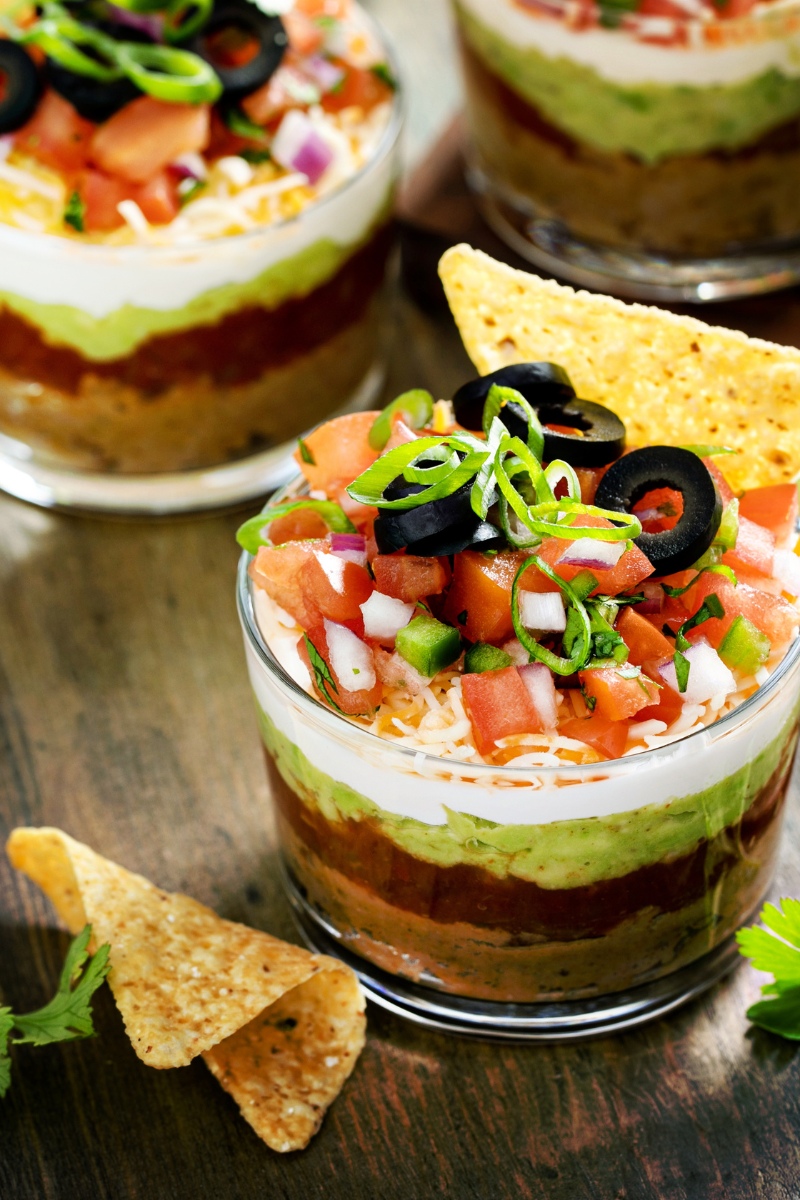 Bowl of layer dip with a chip in it on a wood table in front of another bowl of the same dip