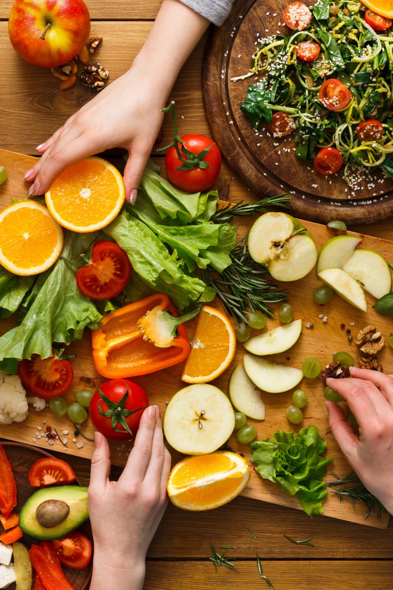 cutting board on a wood table filled with vegetables and fruit with hands grasping for it