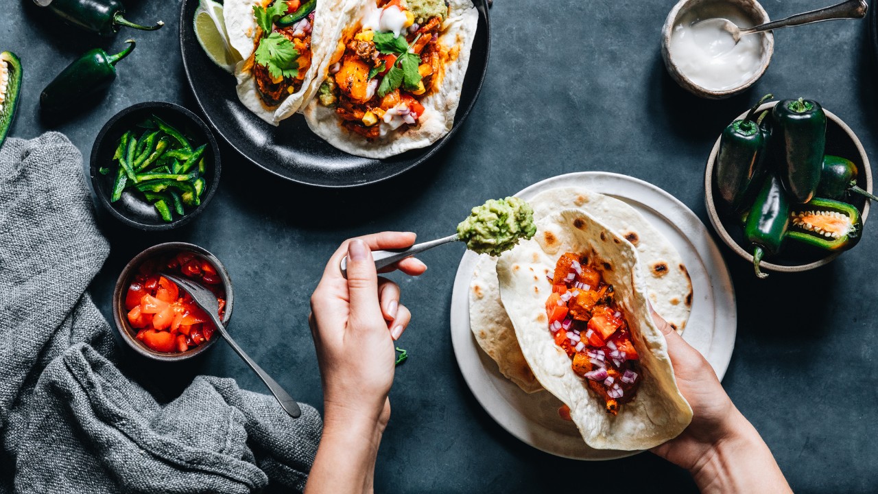 a person holding a taco in one hand over a plate and a fork of guacamole in the other over a dark table next to a plate of tacos