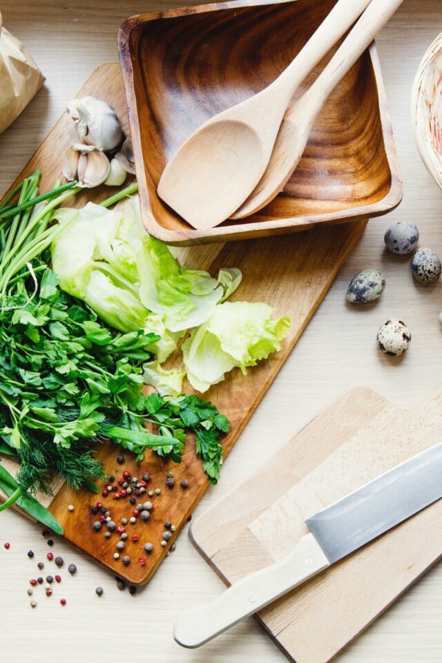 cutting with vegetables on it next to a wood salad bowl with salad tongs inside next to a knife