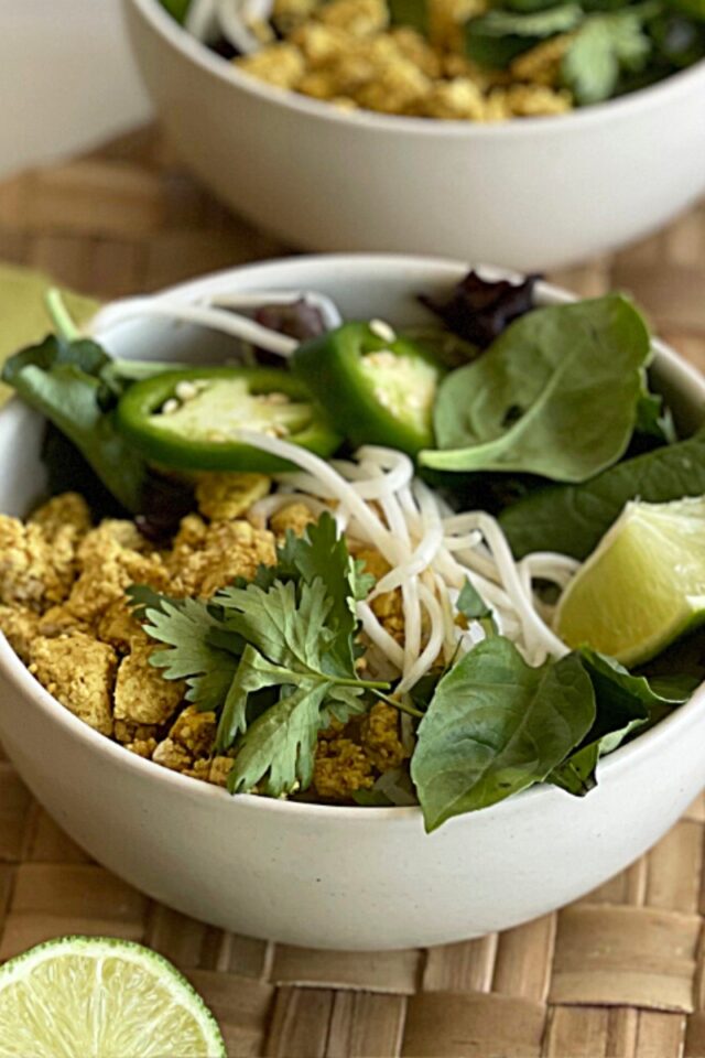 bowl of tofu, bean sprouts, basil leaves, jalapeno and limes on a rattan placemat in front of another bowl