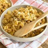 Bowl of pasta salad on an orange and white towel with wooden spoons in it on a wicker placemat