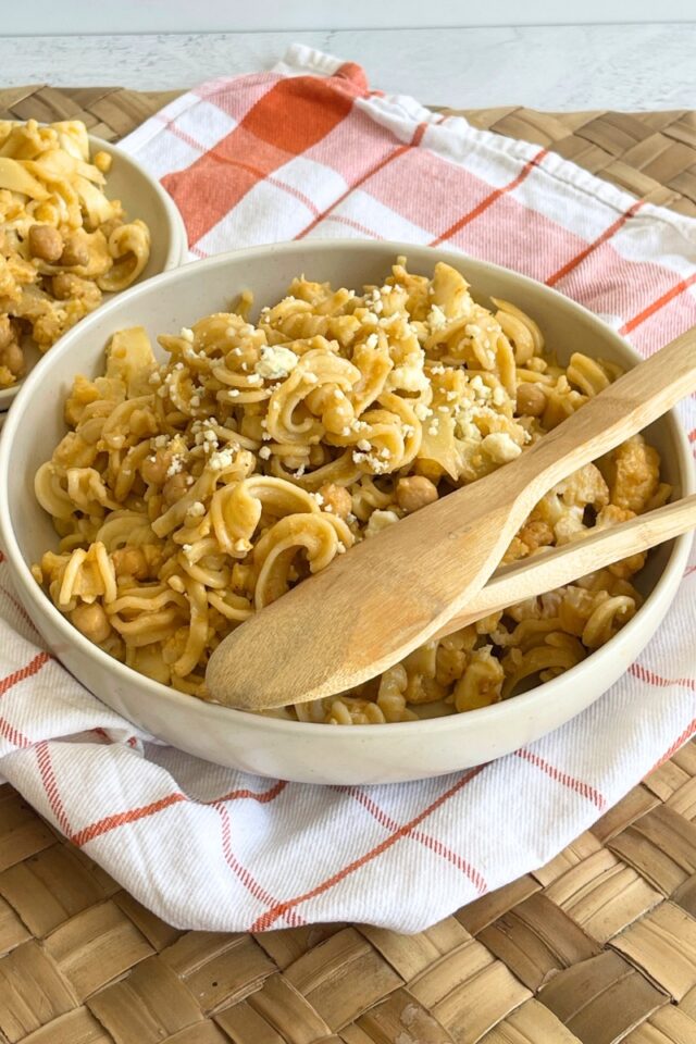 Bowl of pasta salad on an orange and white towel with wooden spoons in it on a wicker placemat