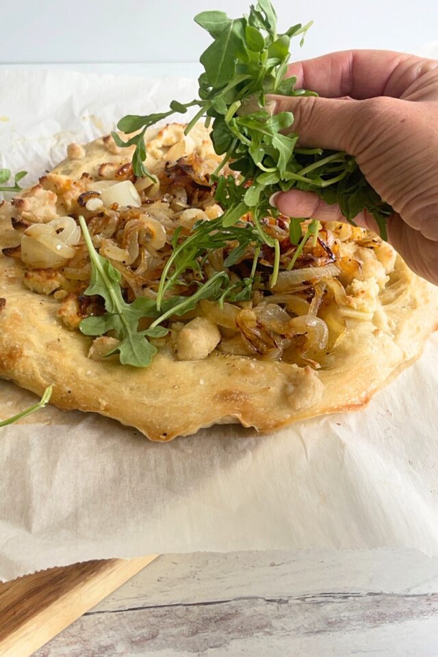 Pizza on parchment paper with a hand sprinkling arugula on top of it