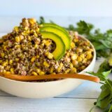 bowl of corn and quinoa salad on a counter next to cilantro and an avocado