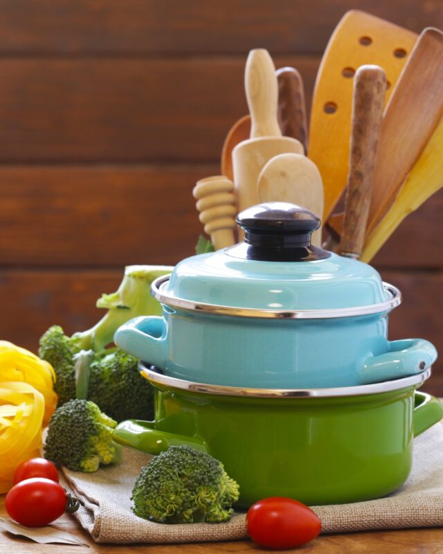 Wooden cooking utensils and a blue pot and a green pot on a kitchen counter alongside broccoli and tomatoes