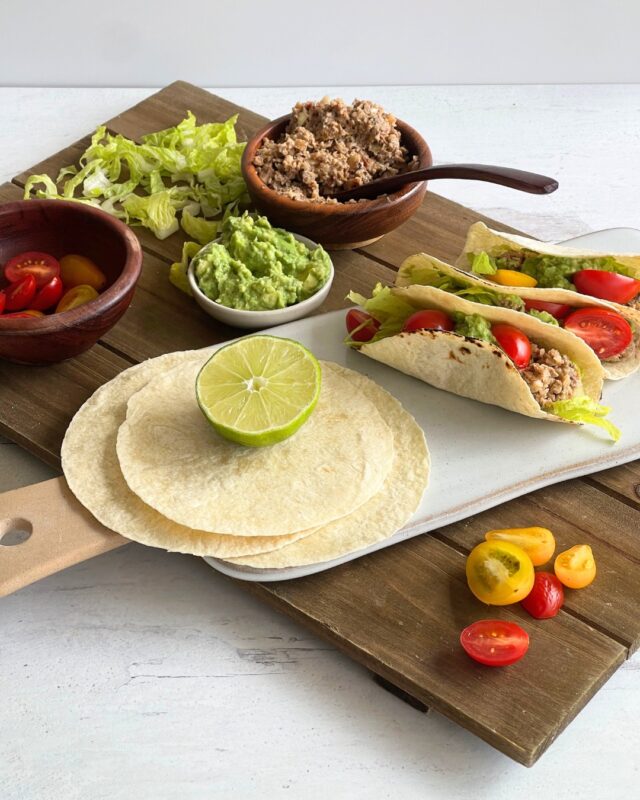 3 tacos on a cutting surrounded by bowls of guacmole and chorizo, and a stack of corn tortillas