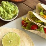 three tacos on a counter next to a wood cutting board, bowl of guacamole and stack of corn tortillas with a lime on top