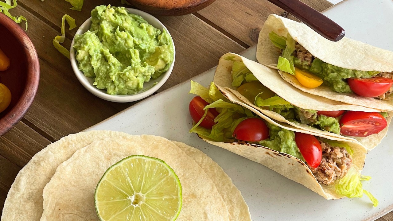 three tacos on a counter next to a wood cutting board, bowl of guacamole and stack of corn tortillas with a lime on top