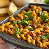 pan of mac and cheese on a checkered cloth in front of a breadboard