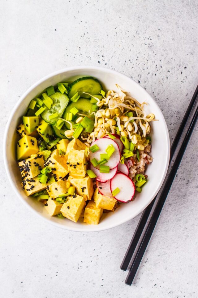 bowl full of tofu, avocado, cucumber and radishes next to chopsticks