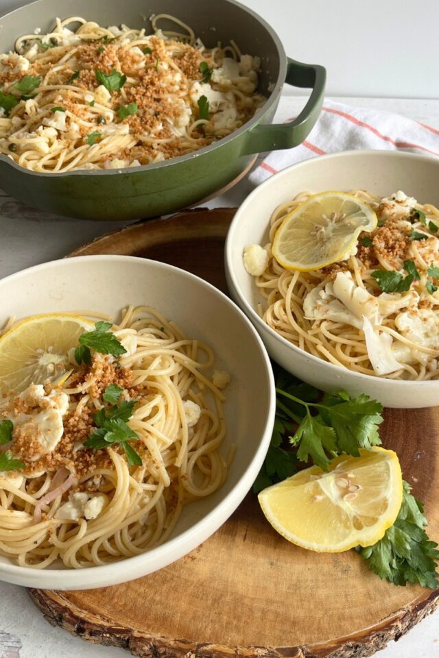 Two bowls of pasta on a wood cutting board in front of a pan of pasta