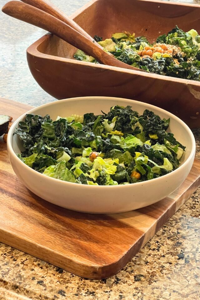 Bowl of salad on wood cutting board in front of a wood salad bowl on the counter