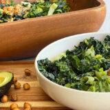 Bowl of salad in front of a wood salad bowl on a cutting board next to a half of avocado