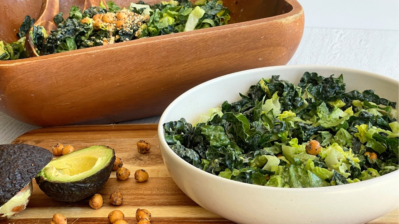 Bowl of salad in front of a wood salad bowl on a cutting board next to a half of avocado