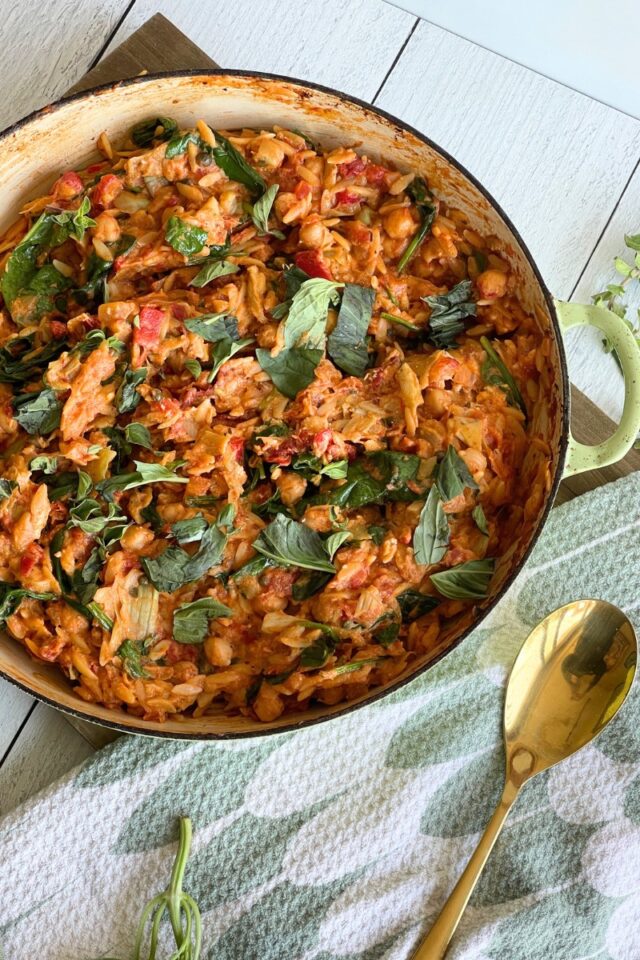 casserole round dish full of tomatoes, orzo and vegetables on a green and white towel alongside a gold spoon