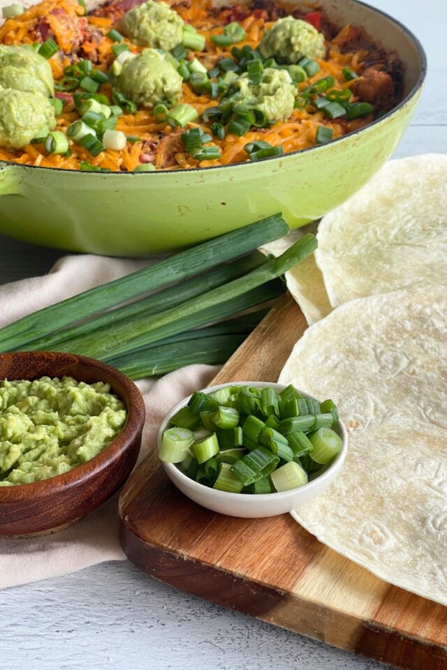 Tortillas and bowls of green onions and guacamole on a wood cutting board in front of a casserole dish of a Mexican rice dish