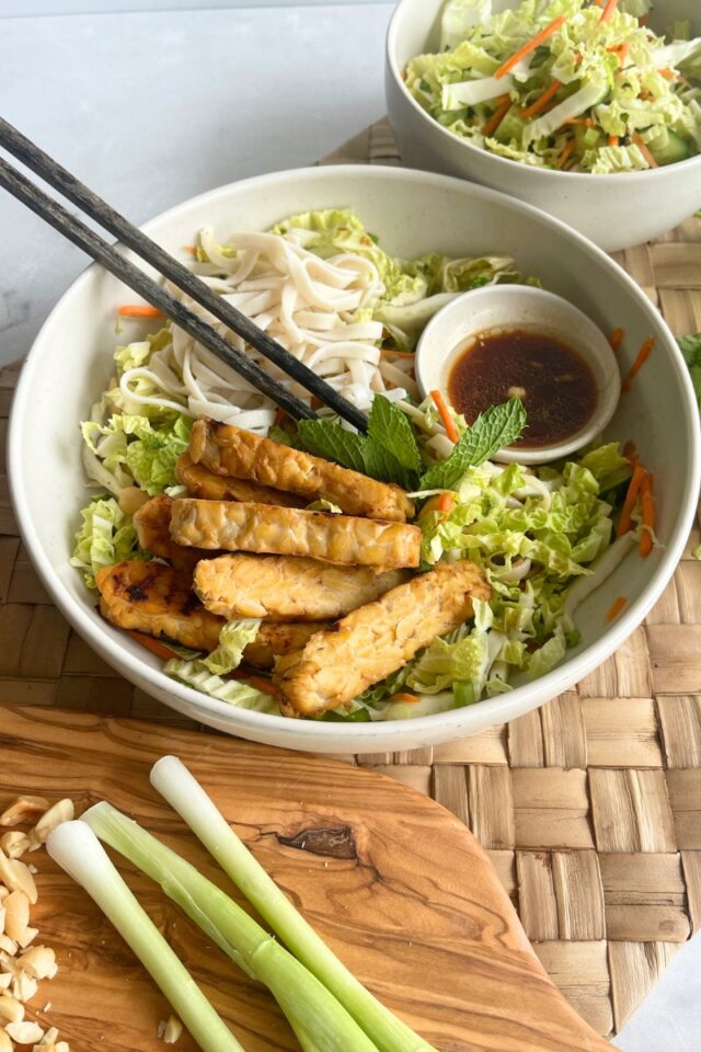 rice noodles, and tempeh in a bowl with chopsticks next to a bowl of cabbage and a cutting board with green onions