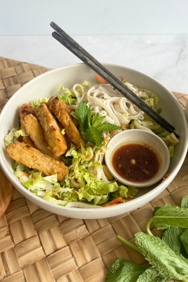 bowl of tempeh, rice noodles and cabbage with chopsticks on a wicker placemat