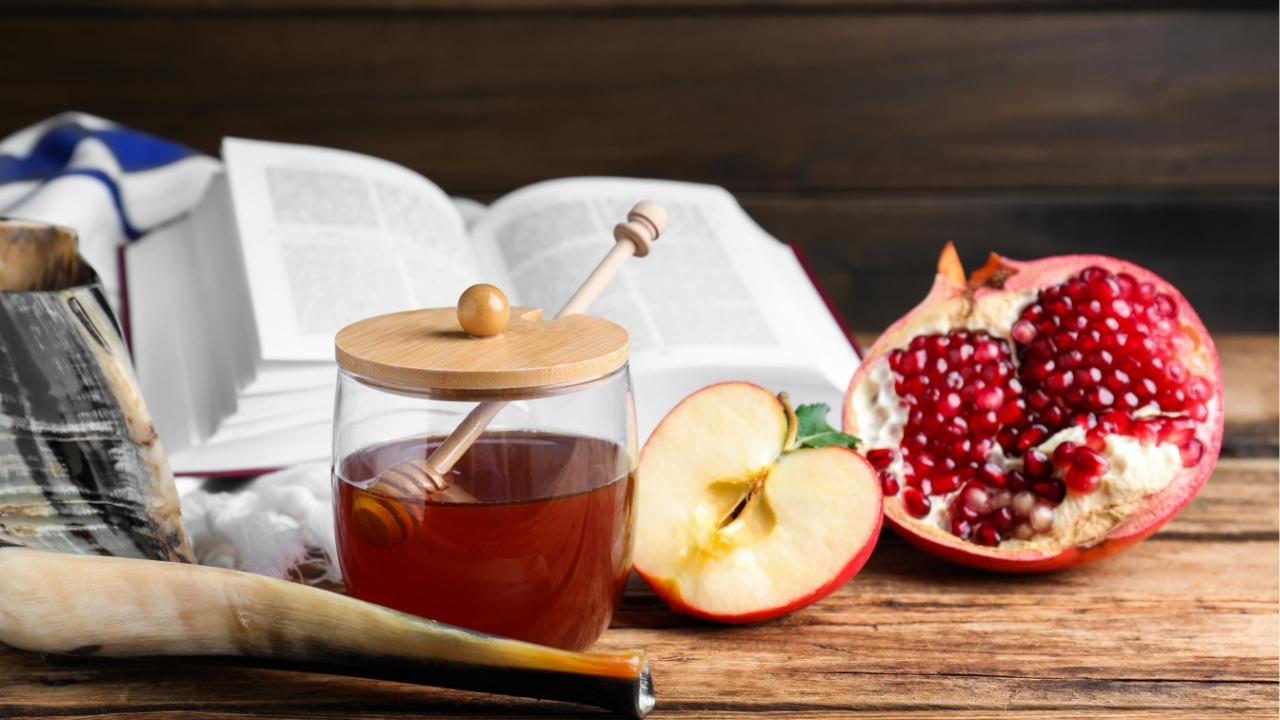 wood table with a jar of honey, a prayer book, half an apple and half a pomegranate