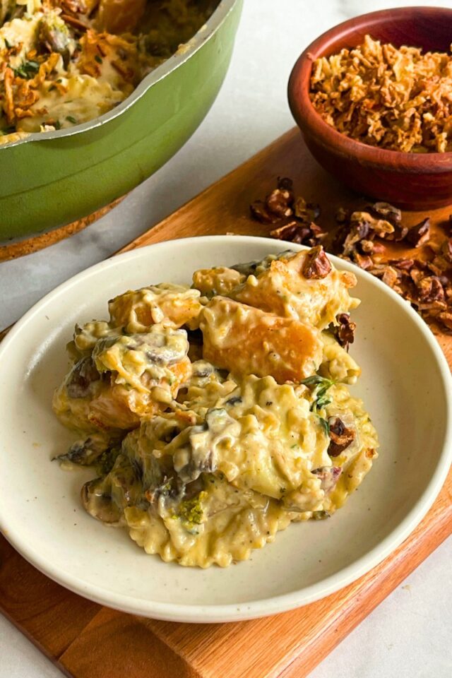 plate with ravioli and butternut squash on it, atop a wood cutting boar next to a small wooden bowl filled with fried onions