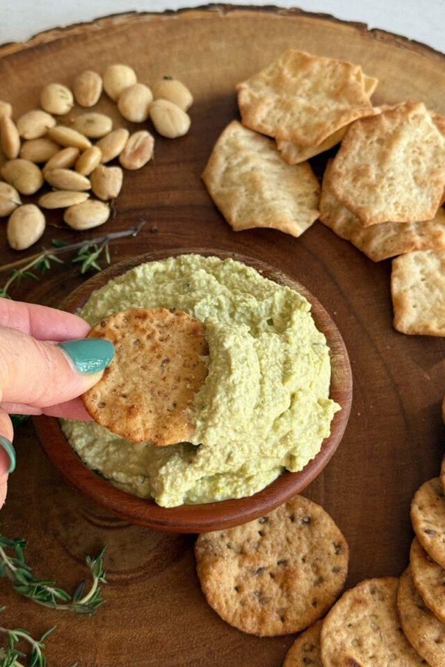 a hand dipping a cracker into edamame dip in a wood bowl on a wooden board surrounded by crackers